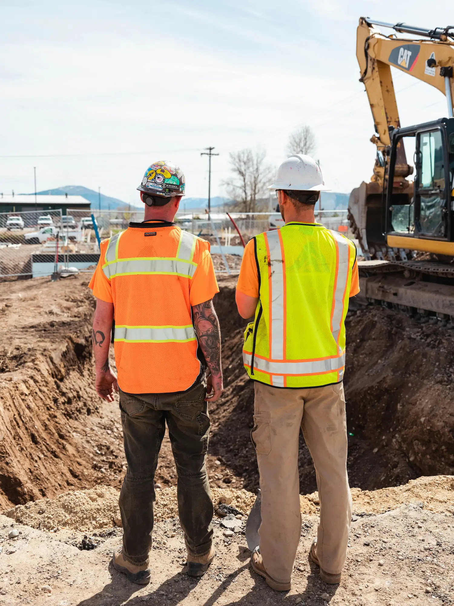 Baggerarbeiten, Erdbeweung: Zwei Männer von hinten, eine Baustelle mit Bagger im Hintergrund.