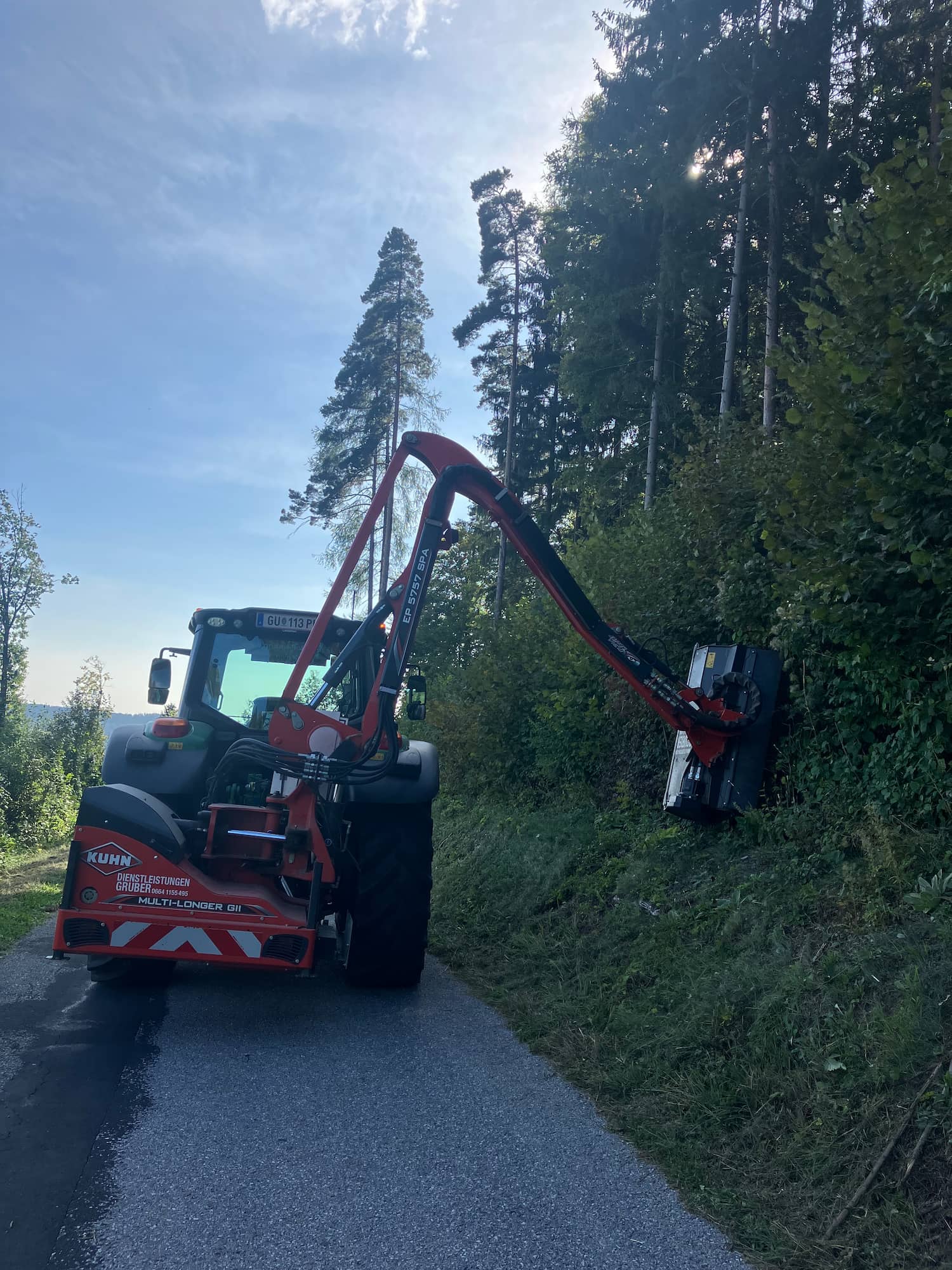 Das Bild zeigt einen Traktor auf einer Straße, der über einen langen Arm die Waldböschung schneidet und trimmt.