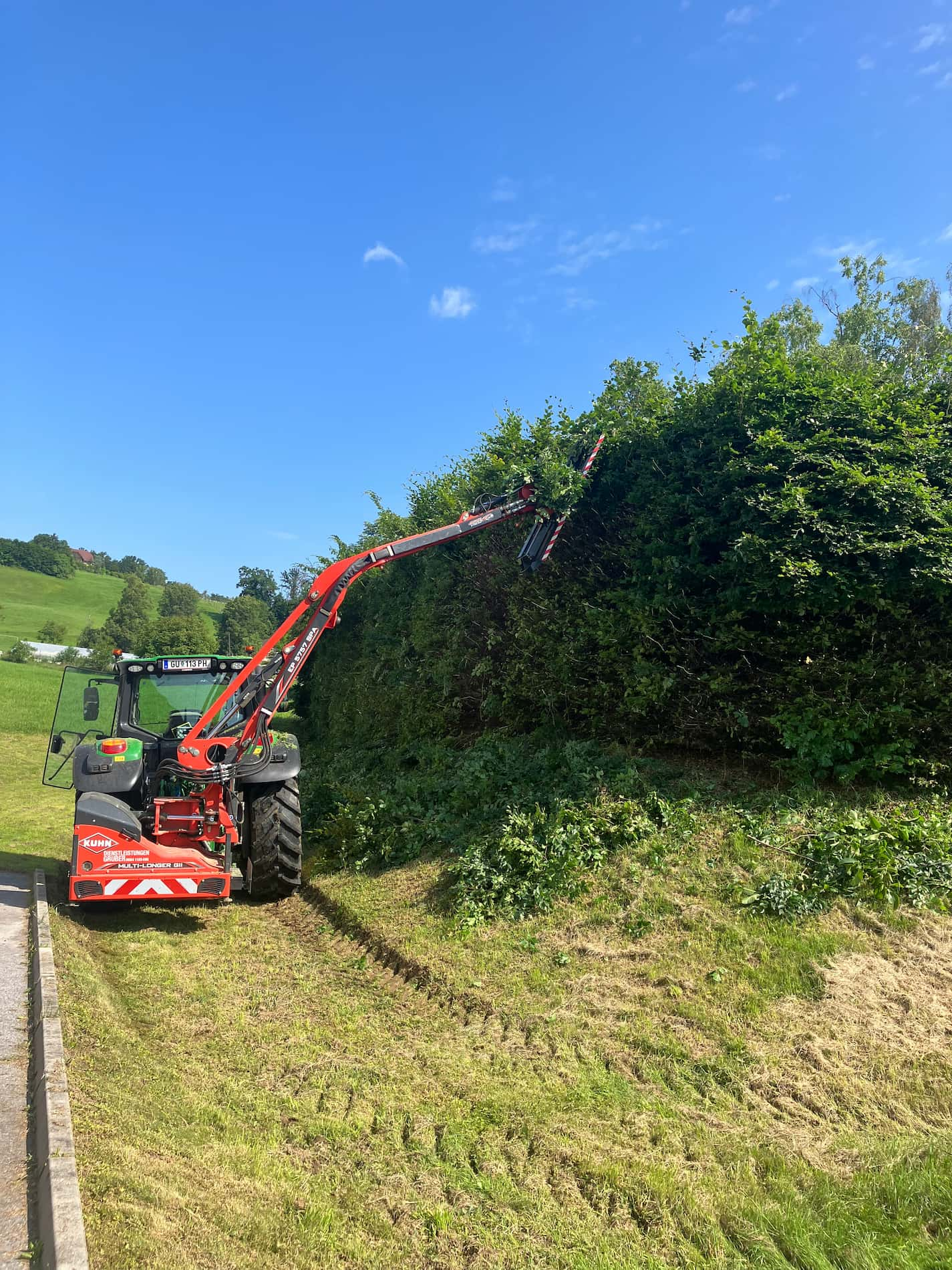 Das Bild zeigt einen Traktor, der auf einer Böschung eine Hecke schneidet.